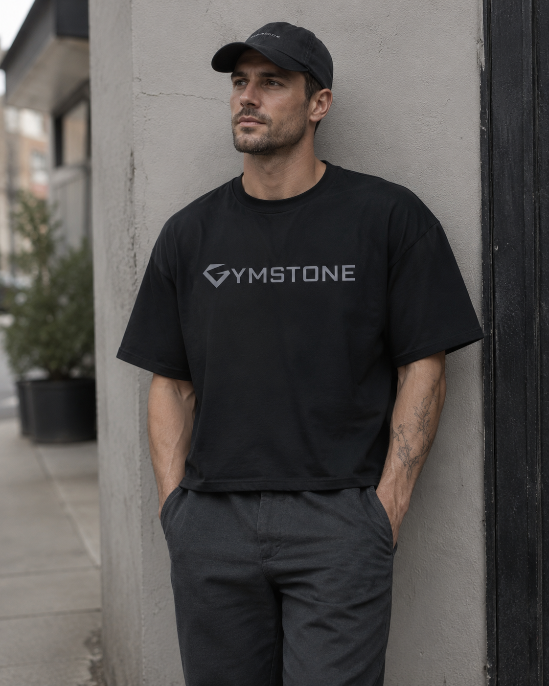 Black - A man wearing a black Gymstone t-shirt and a black baseball cap leans against a concrete pillar in an outdoor urban setting.