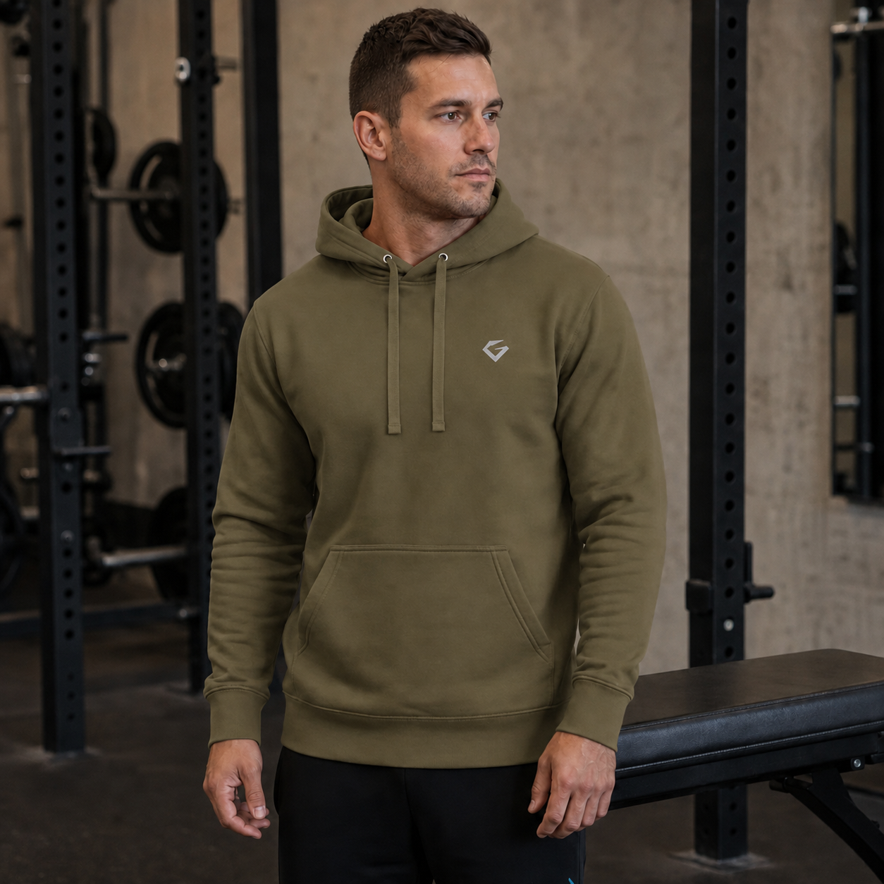 Military Green - A man in a gym wearing an olive green athletic hoodie with a small white geometric logo on the left chest, paired with black sweatpants.