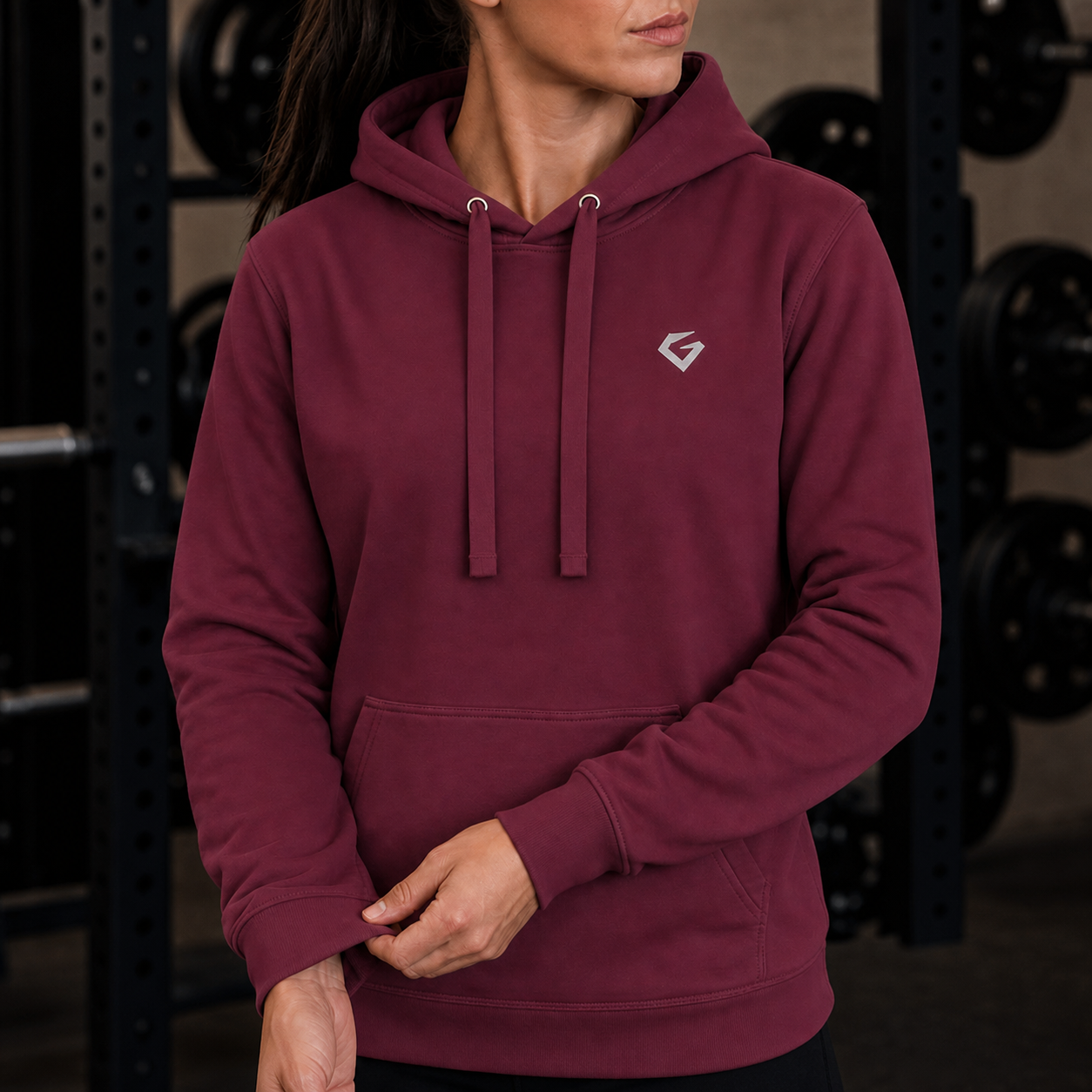 Maroon - A woman wearing a maroon pullover hoodie with a small white geometric logo on the left chest, standing in a gym with weight racks in the background.
